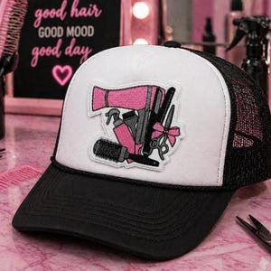 May include: A black and white trucker hat with a patch featuring pink and gray hair styling tools. The hat has a black brim and mesh back. The patch includes a hairdryer, scissors, and a comb. The background includes a sign that says "good hair good mood good day".