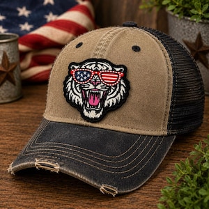 May include: A distressed baseball cap with a tan crown and black mesh back. The cap features a patch of a tiger wearing sunglasses with an American flag design. The bill is black and has distressed edges.