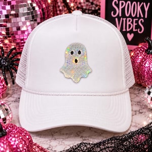 May include: A white trucker hat featuring a central ghost graphic composed of shimmering, iridescent rhinestones. The cap has a structured front panel, a curved brim, a matching white cord detail across the base of the crown, and mesh side panels.