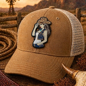 May include: A brown trucker hat with a tan mesh back and a patch featuring a cowgirl in a hat. The hat has a curved brim and a button on top. The background shows a sunset over a rural landscape.