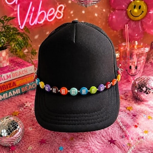 May include: A black trucker hat featuring a decorative chain across the front panel. The chain is composed of small, round, multi-colored beads, each marked with a white letter E, interspersed with tiny silver-toned metallic spacer beads.