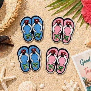 May include: Four embroidered flip-flop patches in blue and pink, each with a red heart and palm tree design. The patches are arranged on a sandy surface with a straw hat, sunglasses, and a postcard that reads "Good Vibes & Tan Lines."