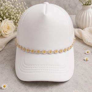 May include: A white baseball cap featuring a mesh back panel and a decorative chain embellishment across the front. The chain consists of a series of small, white daisy flowers with yellow centers that wrap around the crown of the hat.