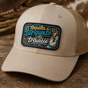 May include: Tan trucker hat with a mesh back and a curved brim. The front features a rectangular patch with the words "tequila turquoise & trouble" and a graphic of a hand holding a cocktail.