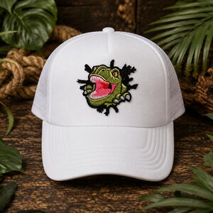 May include: A white trucker hat featuring an embroidered patch of a green dinosaur head with an open mouth and sharp teeth, appearing to burst through a black jagged shape. The cap has a solid front panel and a mesh back for ventilation.