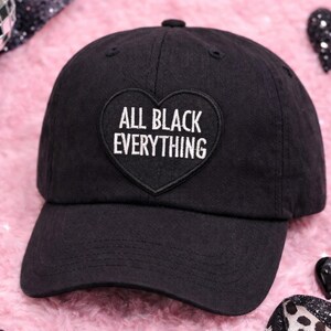 May include: Black baseball cap with a curved brim. The cap features a heart-shaped patch with the white text "ALL BLACK EVERYTHING". The hat is set against a pink, fluffy background with decorative elements.