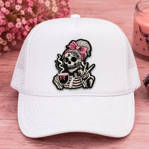May include: A white trucker hat featuring a central graphic patch of a skeleton with a pink bow in its hair, wearing a striped shirt, holding a steaming cup of coffee, and making a peace sign with its hand. The hat has a mesh back and a solid front.