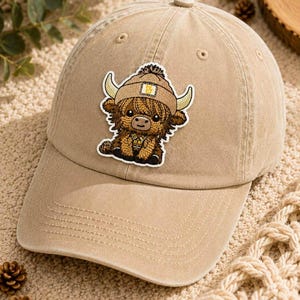 May include: A tan baseball cap featuring a patch of a cartoon Highland cow wearing a hat. The cap has a curved brim and a button on top. The cow patch is brown, tan, and white with yellow horns.