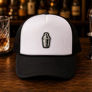 May include: A black and white trucker hat featuring a solid white front panel with a black embroidered cocktail shaker patch, a black brim, and black mesh side and back panels. The hat has a structured crown and a black cord detail above the brim.