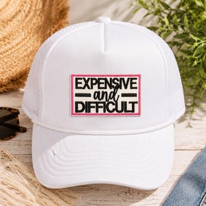 May include: A white trucker hat featuring a rectangular patch on the front panel. The patch has a pink border and contains the black text EXPENSIVE and DIFFICULT with two horizontal lines separating the words on a cream-colored background.
