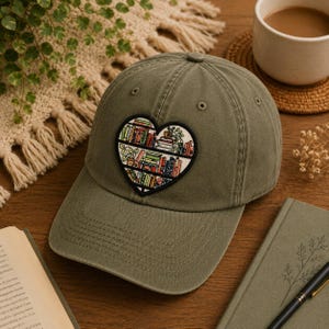 May include: Olive green baseball cap with a heart-shaped patch featuring a bookshelf design. The patch showcases colorful books and plants. The cap is made of a soft material and has a curved brim.