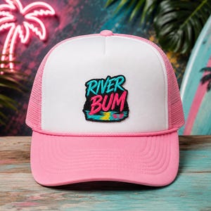 May include: A white and pink trucker hat featuring a central patch with the text RIVER BUM in bright blue and pink lettering. The design includes a stylized sunset over water. The hat has a solid white front panel, pink mesh back, and pink brim.