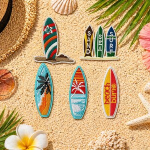 May include: A collection of embroidered surfboard patches with beach-themed designs, including palm trees, sunsets, and the words "Sand", "Sea", "Surf", and "Beach Bum". The patches are arranged on a sandy surface with a straw hat, sunglasses, and tropical flowers.