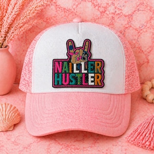 May include: Pink and white trucker hat with a colorful patch that says "NAILLER HUSTLER" and a peace sign hand gesture. The hat has a pink brim and mesh back, perfect for nail technicians or anyone in the beauty industry.