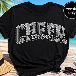 May include: Black t-shirt with the text "CHEER MOM" in white rhinestone-style lettering. The shirt has short sleeves and a crew neck. A black circle with the words "transfer only" is in the upper right corner. Sunglasses and a straw hat are in the background.