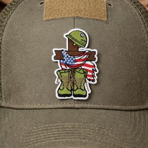 May include: Olive green baseball cap with a mesh back and a patch featuring a scarecrow-like figure in a military helmet, boots, and an American flag. The patch is centered on the front of the cap.
