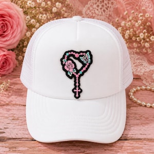 May include: A white trucker hat featuring a central patch design of a rosary shaped into a heart. The patch includes a pink rose, a blue and white butterfly, and a small cross at the bottom, all outlined in black against the white fabric.