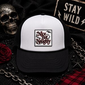 May include: A white and black trucker hat featuring a square patch on the front panel. The patch displays the text Nice But Naughty in a dark red script font, accented by a small black bat silhouette and thorny vine illustrations on a white background.