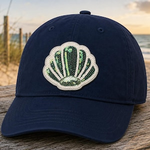 May include: Navy blue baseball cap with a seashell patch. The patch features a green sequined seashell design with a white border. The cap is made of a soft material and has a curved brim.