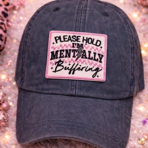 May include: A dark gray baseball cap with a curved brim. The cap features a rectangular patch with the text "PLEASE HOLD I'M MENTALLY Buffering" in black and pink. The hat is made of a soft material.