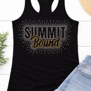 May include: Black racerback tank top with the text "SUMMIT Bound" in white and gold rhinestones. The tank top is paired with denim shorts. A casual outfit for a summer day.