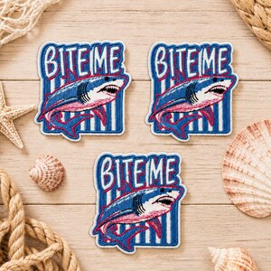May include: Three embroidered patches featuring a blue and white shark with a pink underbelly, swimming over a blue and white vertical striped background. The text BITE ME is embroidered in bold blue letters above the shark on each patch.