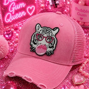 May include: Pink baseball cap with a distressed brim and a mesh back. The cap features an embroidered tiger wearing pink glasses and blowing a bubble. The text "Bubble Gum Queen" is visible in the background.