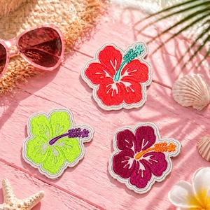 May include: Three embroidered hibiscus flower patches in red, lime green, and magenta, each with a contrasting center and a cream-colored border. The patches are arranged on a pink wooden surface with sunglasses, a straw hat, seashells, and a starfish.