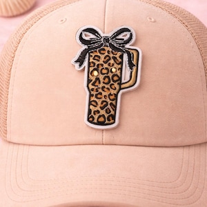 May include: Tan trucker hat with a mesh back and a curved brim. The hat features an embroidered patch of a leopard print tumbler with a black bow. The hat is a neutral color and suitable for various outfits.
