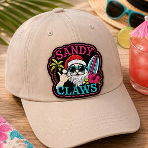 May include: A tan baseball cap with a patch featuring Santa Claus wearing sunglasses and holding a surfboard. The patch reads "SANDY CLAUS" and "CLAWS" in a tropical design. A fun, summer-themed hat.
