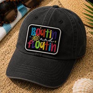 May include: A black baseball cap with a colorful embroidered patch that reads "boatin and floatin". The patch features a sun and various colors. Sunglasses and a beach towel are visible in the background.