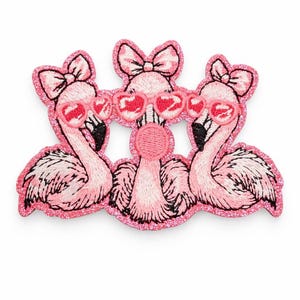 May include: Embroidered patch featuring three pink flamingos wearing heart-shaped sunglasses and bows. The patch has a glittery pink border and is suitable for decorating clothing or accessories.