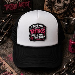 May include: A black and white trucker hat featuring a central patch with the text F-BOMB MOM WITH TATTOOS PRETTY EYES AND THICK THIGHS in pink and white lettering. The cap has a white front panel, a black brim, and black mesh side panels.