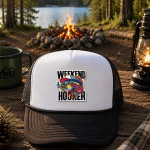 May include: A white and black trucker hat featuring a graphic patch of a colorful fish wearing sunglasses and a hat, with the text WEEKEND HOOKER printed in black block letters above and below the fish illustration on the front panel.