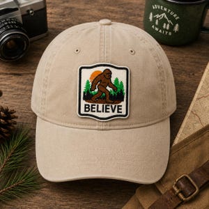 May include: A tan baseball cap with a patch featuring a Bigfoot illustration, a setting sun, and the word "BELIEVE". The cap is made of a soft material and has a curved brim. A vintage camera and a green mug are in the background.