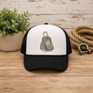 May include: A white and black trucker hat featuring a centered embroidered patch of two grey dog tags. The front panel is white with a black brim and black mesh back. The dog tag design includes a small star detail on the top tag, outlined in a light beige thread, creating a distinct contrast against the white fabric of the hat's front panel.