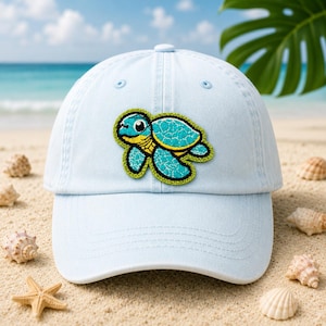 May include: Light blue baseball cap featuring a colorful embroidered sea turtle patch. The turtle has a turquoise shell, yellow accents, and a green outline. The cap is on a sandy beach with seashells and the ocean in the background.