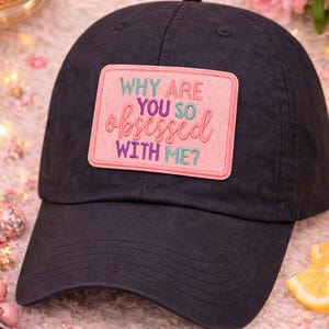May include: A black baseball cap with a rectangular patch that reads "WHY ARE YOU SO OBSESSED WITH ME?" in pink, purple, and green lettering. The cap has a curved brim and a button on top.