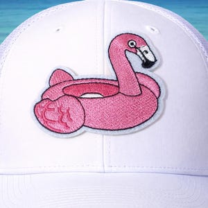 May include: White baseball cap with a pink flamingo float patch. The flamingo is embroidered with black and white details. The cap has a white mesh back and a curved brim, perfect for sun protection.