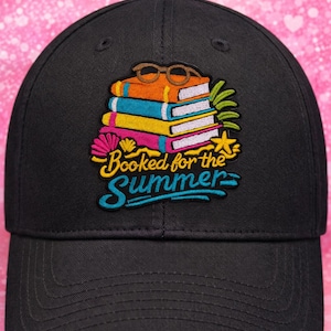 May include: Black baseball cap with an embroidered design featuring a stack of colorful books, eyeglasses, and the text "Booked for the Summer." The design includes a flower and a starfish. The cap has a curved brim.