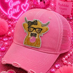 May include: Pink distressed baseball cap with a mesh back and a patch of a cartoon cow wearing glasses and a bow tie, blowing a bubble. The text "Bubble Gum Girl" is displayed on a neon heart-shaped sign.