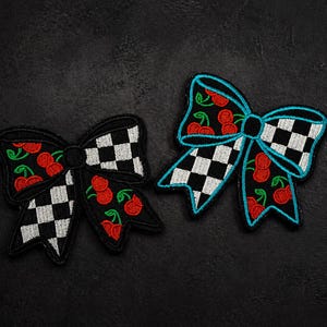 May include: Two embroidered bow patches. One is black with a checkerboard pattern and red cherries. The other bow is black with a checkerboard pattern, red cherries, and a turquoise outline. Both have a black center.