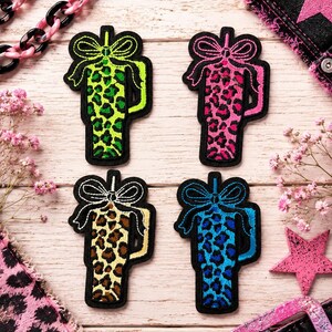 May include: Four embroidered patches shaped like tumblers with straws and bows. Each patch features a distinct leopard print pattern in neon green, bright pink, tan, and vibrant blue, all outlined with a thick black border.