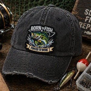 May include: Distressed black baseball cap with a patch that reads "BORN TO FISH FORCED TO WORK" featuring a green and yellow fish. Fishing gear, including a reel, tackle box, and lure, are in the background.