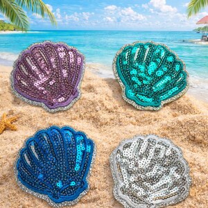May include: Four sequined seashell patches in purple, teal, blue, and silver, with silver trim. The patches are arranged on a sandy beach with a blue ocean and sky in the background. Palm trees and a beach umbrella are visible.