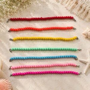May include: Seven beaded strands arranged horizontally, each featuring a single row of matte, round beads in distinct solid colors: red, orange, yellow, mint green, light blue, light pink, and magenta, finished with silver-toned metal lobster clasps.