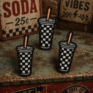 May include: Three embroidered patches shaped like soda cups with black and white checkered patterns. Each patch has a black border and an orange straw. The background includes vintage signs that read "Ice Cold Soda 25¢", "Good Vibes", and "Fast Drinks".