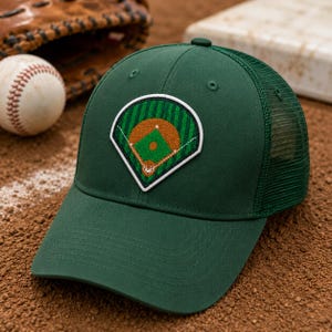 May include: A dark green baseball cap with a mesh back and a baseball field patch. The patch features a brown infield, green outfield, and white outline. A baseball and glove are in the background.