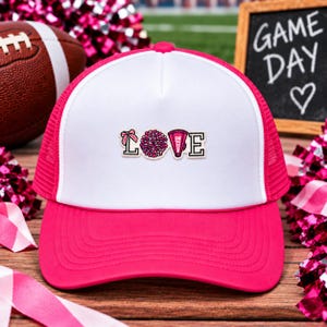 May include: A white and hot pink trucker hat featuring a graphic on the front panel that spells the word LOVE. The letter L is decorated with a pink bow, the letter O is a pink pom-pom, the letter V is a pink megaphone, and the letter E is plain.