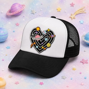 May include: A black and white trucker hat featuring a central embroidered patch of a black heart shape filled with white concentric lines and colorful celestial bodies including planets, stars, and a comet. The hat has a solid black brim and mesh back.